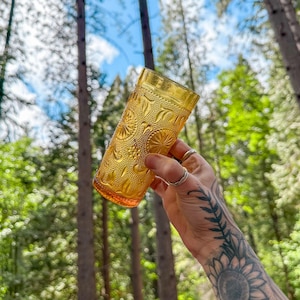 May include: A vintage amber-colored glass with a detailed embossed pattern of circles and floral designs. The tall, cylindrical glass is held up against a backdrop of green trees and blue sky.