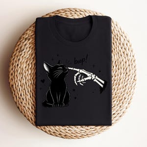 May include: A black t-shirt featuring a graphic print of a black cat sitting and being touched on the nose by a skeletal finger. The text "boop!" is printed above the cat, accompanied by small star and heart icons surrounding the illustration.