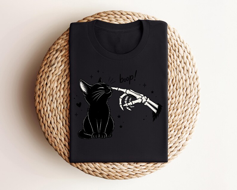 May include: A black t-shirt featuring a graphic print of a black cat sitting and being touched on the nose by a skeletal finger. The text "boop!" is printed above the cat, accompanied by small star and heart icons surrounding the illustration.