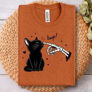 May include: A burnt orange t-shirt featuring a graphic of a black cat being touched on the nose by a skeletal finger. The text "boop!" is printed above the cat, accompanied by small star and heart illustrations. The shirt has a crew neckline.