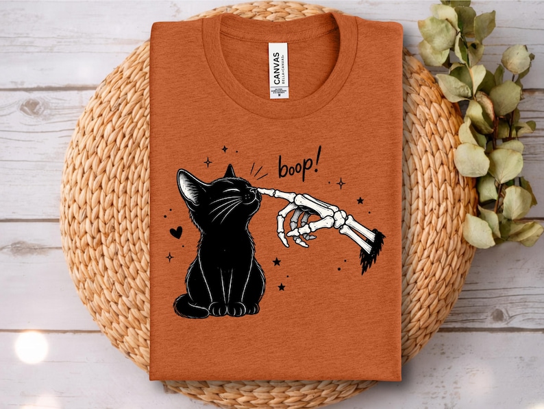 May include: A burnt orange t-shirt featuring a graphic of a black cat being touched on the nose by a skeletal finger. The text "boop!" is printed above the cat, accompanied by small star and heart illustrations. The shirt has a crew neckline.