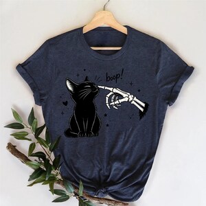 May include: A dark heather navy blue short-sleeve t-shirt featuring a graphic print of a black cat being touched on the nose by a skeletal finger. The text "boop!" is printed in a handwritten style above the cat, surrounded by small star icons.