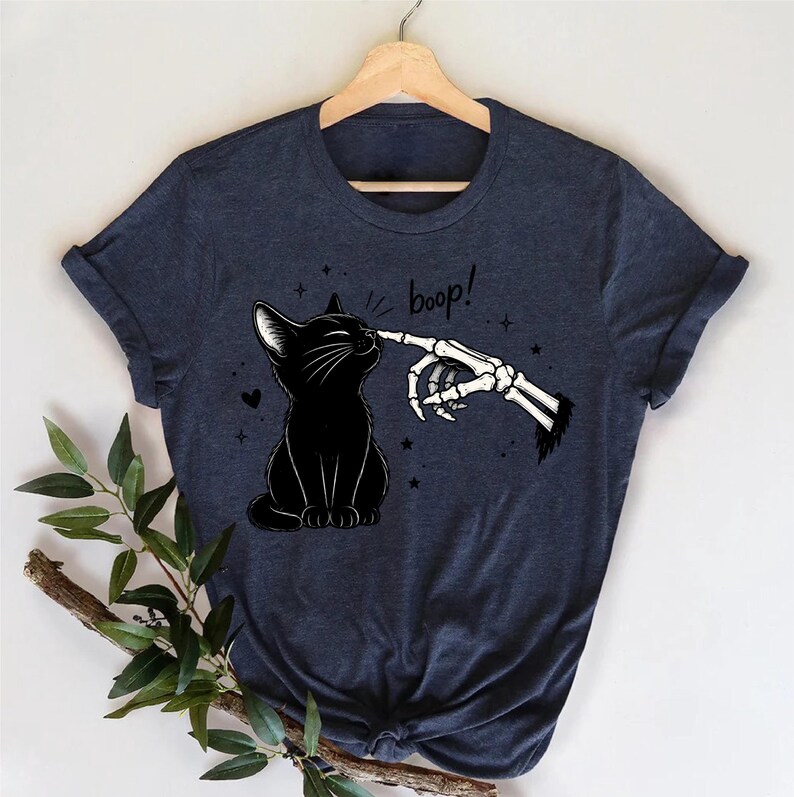 May include: A dark heather navy blue short-sleeve t-shirt featuring a graphic print of a black cat being touched on the nose by a skeletal finger. The text "boop!" is printed in a handwritten style above the cat, surrounded by small star icons.