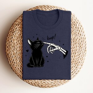 May include: A dark navy blue t-shirt featuring a graphic illustration of a black cat being touched on the nose by a skeletal finger. The text "boop!" is printed above the cat, surrounded by small stars and a heart, creating a detailed design.