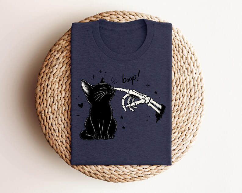 May include: A dark navy blue t-shirt featuring a graphic illustration of a black cat being touched on the nose by a skeletal finger. The text "boop!" is printed above the cat, surrounded by small stars and a heart, creating a detailed design.