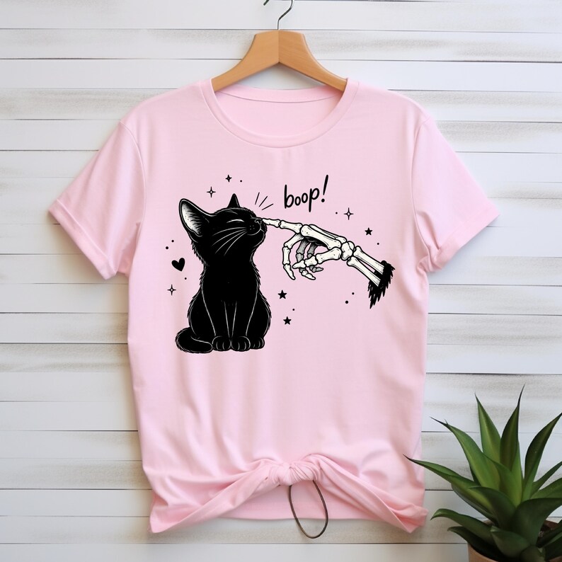 May include: A light pink short-sleeve t-shirt featuring a black graphic of a cat being touched on the nose by a skeletal finger. The design includes the text "boop!" in a black script font, surrounded by small stars and a heart, printed on the front.