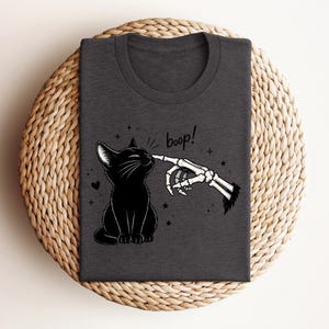 May include: A dark charcoal gray t-shirt featuring a graphic print of a black cat sitting and being touched on the nose by a skeletal finger. The text "boop!" is written in a black script font above the cat, surrounded by small stars and a heart.