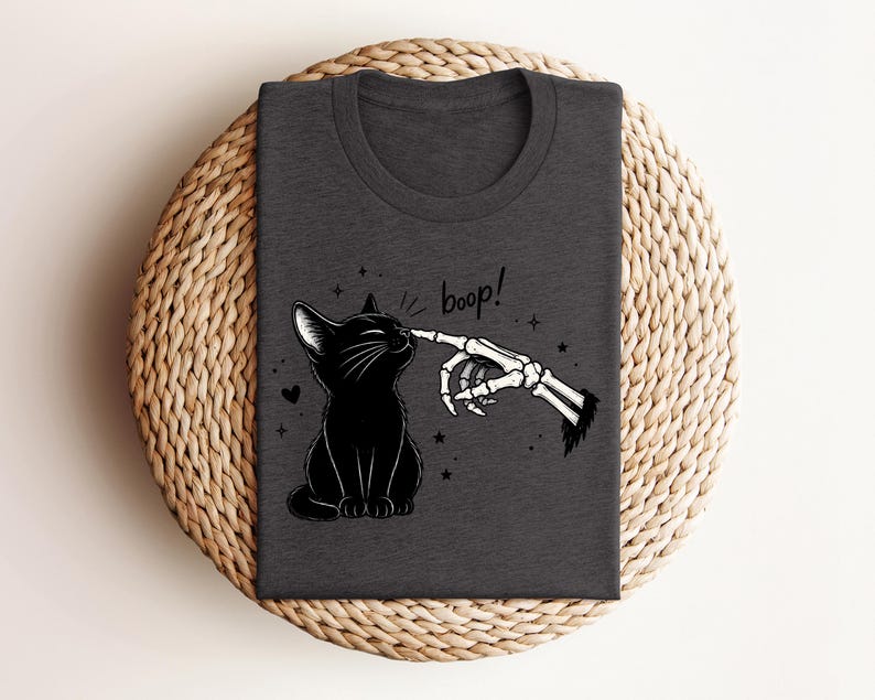 May include: A dark charcoal gray t-shirt featuring a graphic print of a black cat sitting and being touched on the nose by a skeletal finger. The text "boop!" is written in a black script font above the cat, surrounded by small stars and a heart.