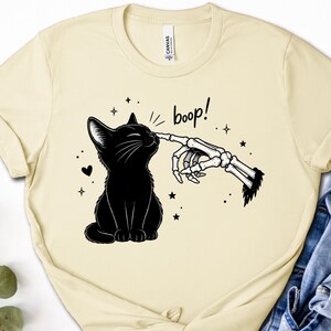 May include: A cream-colored t-shirt featuring a black graphic of a sitting cat being touched on the nose by a skeletal finger. The text "boop!" is printed above the cat, surrounded by small star and heart icons. The shirt has a crew neckline.