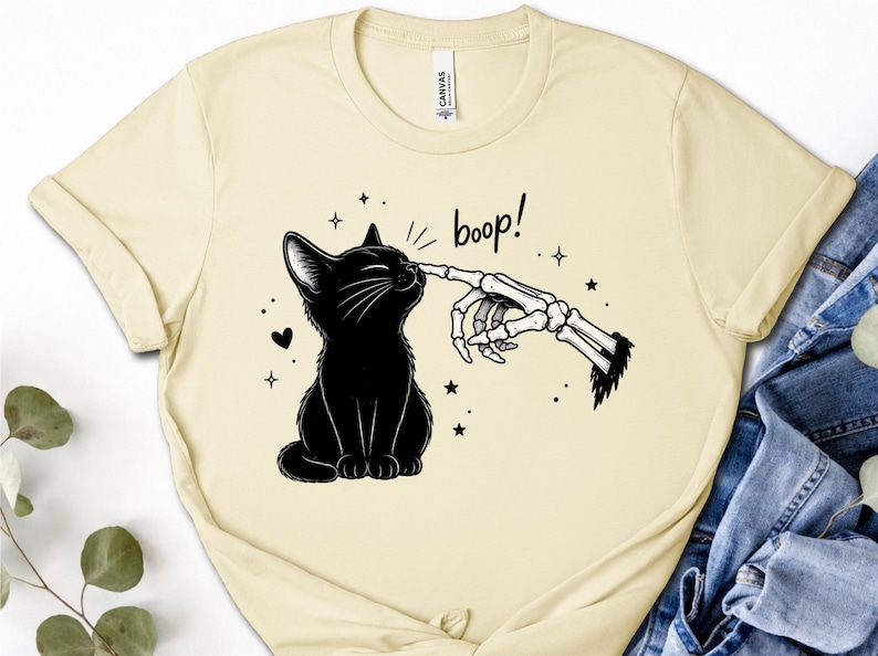 May include: A cream-colored t-shirt featuring a black graphic of a sitting cat being touched on the nose by a skeletal finger. The text "boop!" is printed above the cat, surrounded by small star and heart icons. The shirt has a crew neckline.