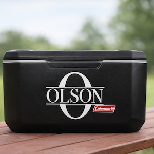May include: A black Coleman cooler featuring a large white letter O with the name OLSON printed in white across the center. The cooler has a rectangular shape with a black lid and a small red and white Coleman logo located in the bottom right corner.