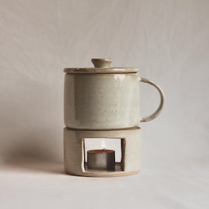 May include: A ceramic mug with a lid resting on a matching cylindrical warmer base. The off-white, speckled glaze covers both pieces. A small, lit tea light candle is visible inside the open rectangular cutout of the warmer base.