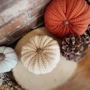 May include: Three crocheted pumpkins in varying sizes and colors, including one large burnt orange pumpkin, one medium beige pumpkin, and one small off-white pumpkin, each featuring a textured ribbed surface and a small brown stem at the center.