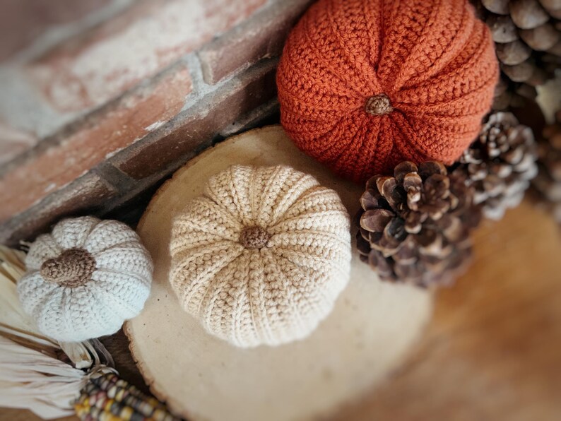 May include: Three crocheted pumpkins in varying sizes and colors, including one large burnt orange pumpkin, one medium beige pumpkin, and one small off-white pumpkin, each featuring a textured ribbed surface and a small brown stem at the center.