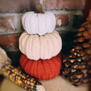May include: A stack of three crocheted pumpkins in varying sizes and colors. The top pumpkin is white, the middle is beige, and the bottom is rust orange. Each pumpkin features a ribbed texture and a small brown crocheted stem at the very top.