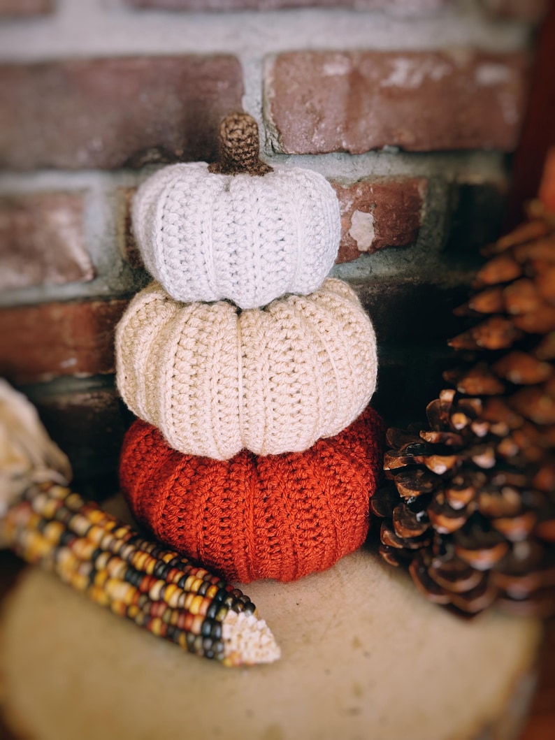 May include: A stack of three crocheted pumpkins in varying sizes and colors. The top pumpkin is white, the middle is beige, and the bottom is rust orange. Each pumpkin features a ribbed texture and a small brown crocheted stem at the very top.