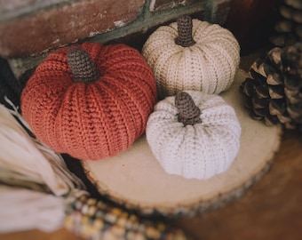 May include: Three crocheted pumpkins in rust orange, cream, and white, each featuring a dark brown stem. The pumpkins are crafted with a ribbed texture and are arranged together, showcasing their rounded shapes and distinct color variations.