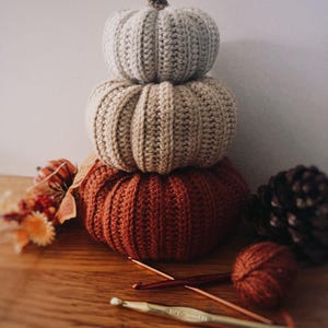May include: A stack of three crocheted pumpkins in varying sizes and colors, featuring a light gray top pumpkin, a beige middle pumpkin, and a large rust-colored bottom pumpkin, each with a textured ribbed pattern and a small brown stem.