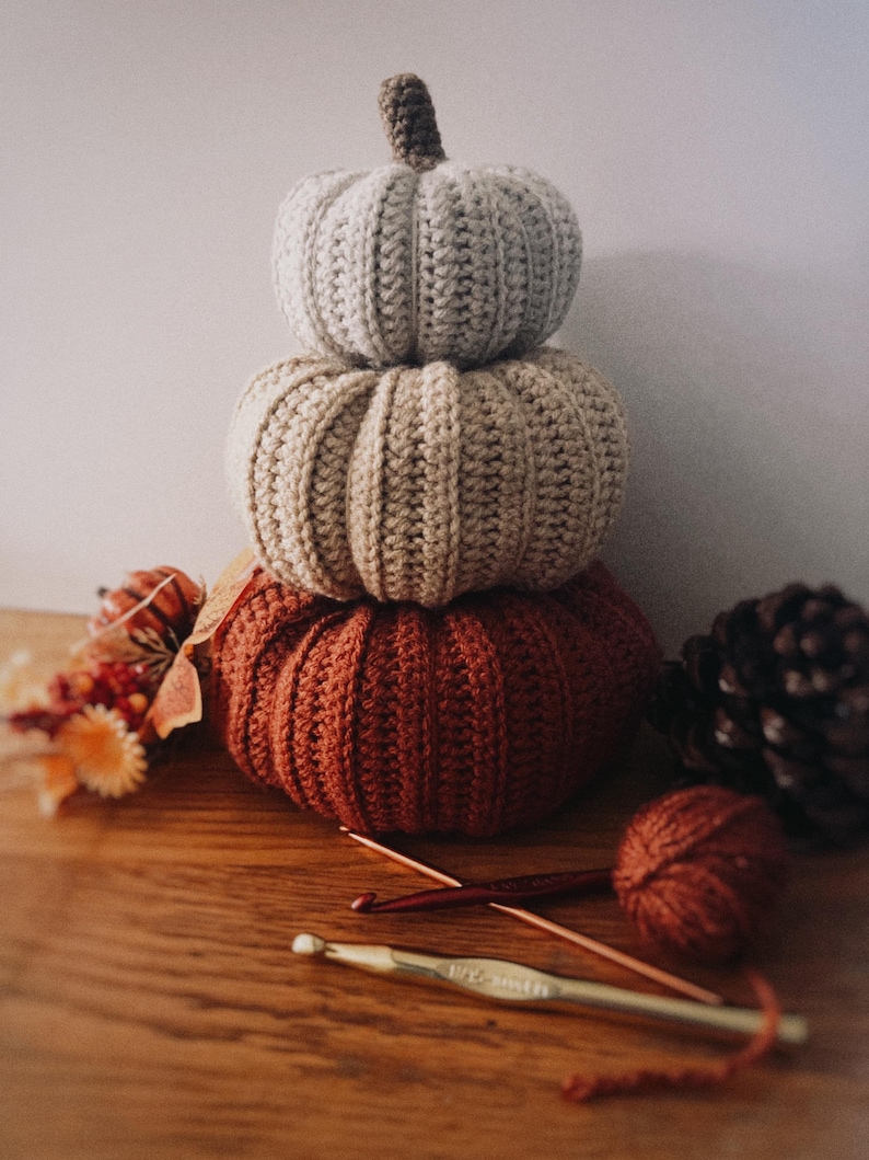 May include: A stack of three crocheted pumpkins in varying sizes and colors, featuring a light gray top pumpkin, a beige middle pumpkin, and a large rust-colored bottom pumpkin, each with a textured ribbed pattern and a small brown stem.