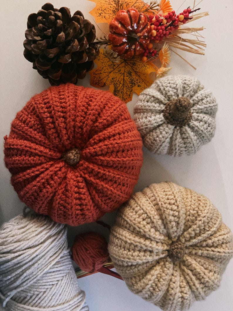 May include: Three crocheted pumpkins in rust, beige, and cream colors with brown stems, accompanied by a large pinecone, a small decorative orange pumpkin, artificial autumn leaves, red berries, and two balls of yarn in cream and rust colors.