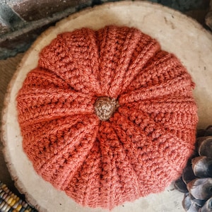 May include: A round, crocheted pumpkin in a burnt orange color with a textured, ribbed surface and a small, brown, tightly crocheted stem at the center. The pumpkin is crafted with visible yarn stitches and features a segmented, rounded shape.