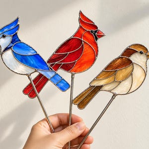 Puede incluir: Tres adornos de aves de vidrio tintado sobre varillas de metal. El primero es un arrendajo azul y blanco, el segundo es un cardenal rojo brillante y el tercero es un gorrión marrón y crema. Cada ave presenta segmentos de vidrio distintos.
