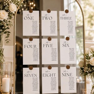 May include: An arched gold-framed mirror displaying nine white rectangular seating chart cards arranged in three rows. Each card features a table number from one to nine, a list of names, and a brown wax seal at the top center of each paper card.
