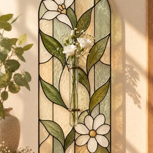 May include: An arched stained glass wall hanging featuring a floral motif with white lilies and daisies, green leaves, and pale yellow panels. A clear glass test tube vase is mounted in the center, holding a small bouquet of white flowers.