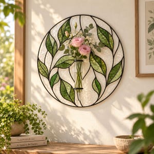 Puede incluir: Una pieza de arte mural circular con un diseño de vidrieras de hojas verdes sobre un fondo blanco texturizado. Un florero cilíndrico de vidrio transparente está fijado en el centro, conteniendo flores rosas y tallos de eucalipto.