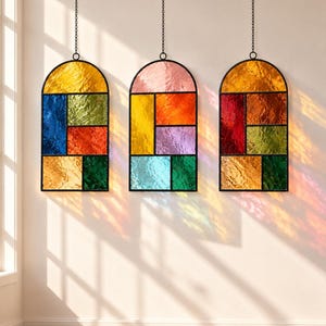 May include: Three arched stained glass panels hanging from black chains. Each panel features a grid of multicolored, textured glass segments in shades of yellow, blue, green, red, orange, pink, and purple, framed in black metal.