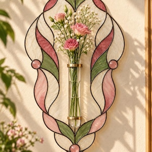 May include: A wall-mounted stained glass panel featuring a central clear glass tube vase. The panel has a decorative metal frame with pink and green glass segments. A bouquet of pink flowers and white baby's breath is inside the glass tube.