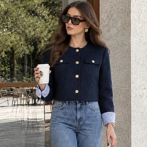 May include: A navy blue cropped jacket with gold buttons and two chest pockets, worn over a blue and white striped long-sleeve shirt. The outfit includes high-waisted wide-leg blue denim jeans, a cream-colored structured handbag, and sunglasses.
