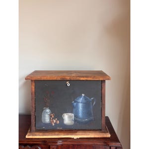 May include: A rectangular wooden bread box with a dark, painted front panel featuring a blue coffee pot, a white mug, a small teddy bear, and a vase with red flowers. The box has a natural wood frame and a small white knob on the front door.