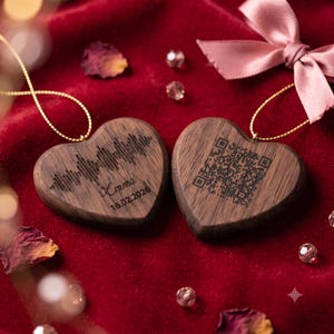 Puede incluir: Dos adornos de madera en forma de corazón con cordones dorados. Uno presenta una onda sonora grabada, el nombre Emma y la fecha 16.02.2026. El otro adorno muestra un código QR grabado. Ambos objetos están hechos de madera de color marrón oscuro.