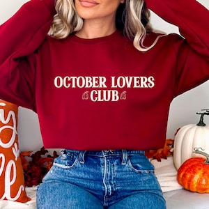 Könnte beinhalten: Ein dunkelrotes Langarm-Sweatshirt mit Rundhalsausschnitt und dem weißen Schriftzug "OCTOBER LOVERS CLUB" auf der Brust, flankiert von zwei kleinen weißen Kürbis-Symbolen. Das Kleidungsstück wird mit einer blauen Jeans mit sichtbaren Abnutzungen kombiniert.