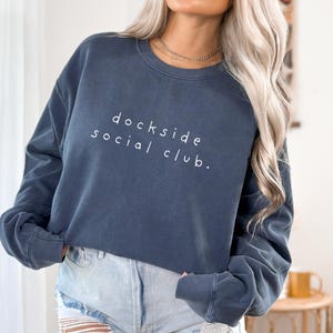 May include: A slate blue long-sleeve crewneck sweatshirt featuring the white text "dockside social club." printed in a simple sans-serif font across the chest. The garment has a relaxed fit with ribbed cuffs and a ribbed neckline.