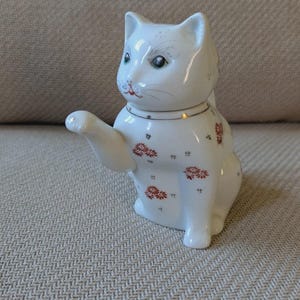 Peut inclure: Une figurine de chat en céramique blanche avec une patte levée, ornée d'un motif délicat de petites fleurs rouges et de détails dorés autour du cou. Le chat présente des traits peints incluant des yeux verts, un nez rose et des moustaches.