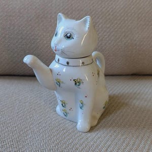 Peut inclure: Une théière en céramique blanche en forme de chat assis. La théière présente un collier peint avec des points dorés, de petits motifs floraux bleus et jaunes sur le corps, et un bec verseur partant de la poitrine du chat. Visage peint.