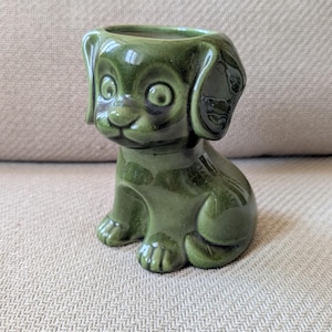 Può includere: Un vaso in ceramica verde oliva lucido a forma di cucciolo seduto con grandi orecchie pendenti, occhi spalancati e un muso arrotondato. La parte superiore della testa del cucciolo presenta un'apertura circolare per piccole piante.