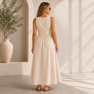 May include: A sleeveless, floor-length cream-colored dress with a fitted bodice, a defined waistline, and a full, flowing skirt. The dress features a simple round neckline and a clean, solid-colored design that reaches down to the ankles.