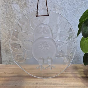 Op de afbeelding: Een ronde wandhanger van helder glas met een centraal geplaatste, gematteerde uilfiguur omringd door reliëfpatronen van bladeren. De glazen schijf heeft een gladde, doorschijnende afwerking en hangt aan een dun bruin leren koord aan de bovenkant.