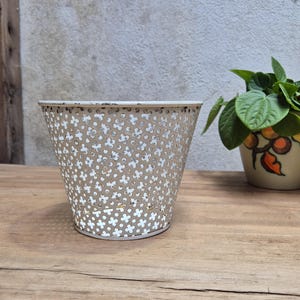 May include: A light beige metal planter with a perforated pattern of small four-petaled flowers. The cylindrical container features a slightly wider top rim and a solid base, showcasing a weathered finish along the upper edge of the metal structure.