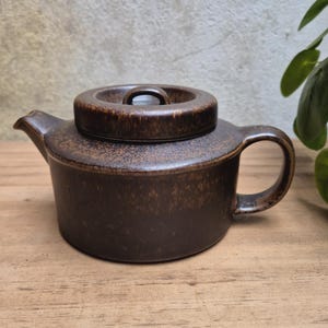 Könnte beinhalten: Eine dunkelbraune Keramik-Teekanne mit gesprenkelter Oberfläche, bestehend aus einem zylindrischen Körper, einem kurzen Ausguss, einem geschwungenen Henkel und einem passenden runden Deckel mit kleinem Knauf. Die Oberfläche ist meliert.