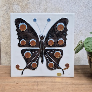 May include: A square white ceramic wall tile featuring a raised, dark brown butterfly with orange and light blue circular accents on its wings. The butterfly has a textured body and two antennae with blue tips. A small gold sticker is in the corner.
