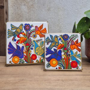 Könnte beinhalten: Zwei quadratische Keramikfliesen in goldfarbenen Metallrahmen, die jeweils eine lebendige, farbenfrohe Illustration stilisierter Vögel und Blumen zeigen. Das Kunstwerk zeigt ein wiederkehrendes Muster aus blauen, orangen und violetten Vögeln.