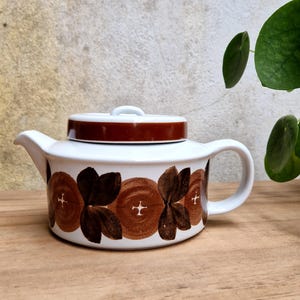 Puede incluir: Una tetera de cerámica blanca con tapa marrón y un patrón repetitivo de formas circulares de color marrón oscuro y motivos de hojas alrededor del cuerpo. La tetera tiene un asa curva, un pico corto y una base plana, con pequeñas cruces blancas en el centro.