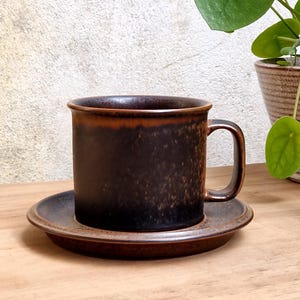 Könnte beinhalten: Ein dunkelbraunes Keramik-Tassenset mit passender Untertasse, das eine gesprenkelte Glasur mit einem helleren braunen Rand aufweist. Die Tasse hat einen zylindrischen Körper und einen C-förmigen Henkel, während die Untertasse einen leicht erhöhten Rand hat.