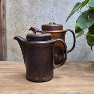 Op de afbeelding: Twee keramische theepotten met deksels, voorzien van een donkerbruin en amberkleurig glazuur met een gespikkelde, getextureerde afwerking. Elke theepot heeft een cilindrisch lichaam, een gebogen handvat en een korte tuit. Ze staan achter elkaar.