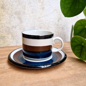 Op de afbeelding: Een keramieken koffiekopje met bijpassende schotel, voorzien van horizontale strepen in zwart, wit, bruin en donkerblauw. Het kopje heeft een witte binnenkant en een gebogen oortje, terwijl de schotel concentrische ringen in dezelfde kleuren toont.