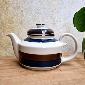 Puede incluir: Una tetera de cerámica con base blanca brillante y rayas horizontales en azul oscuro y marrón. La tetera incluye una tapa a juego con un pequeño pomo, un asa curva y un pico vertedor, todo acabado con un esmalte suave y reflectante.